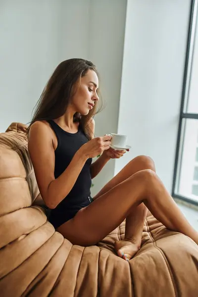 Hübsche junge Frau mit brünetten langen Haaren, die eine Tasse Morgenkaffee in der Hand hält und auf einem Bohnensackstuhl sitzt — Stockfoto