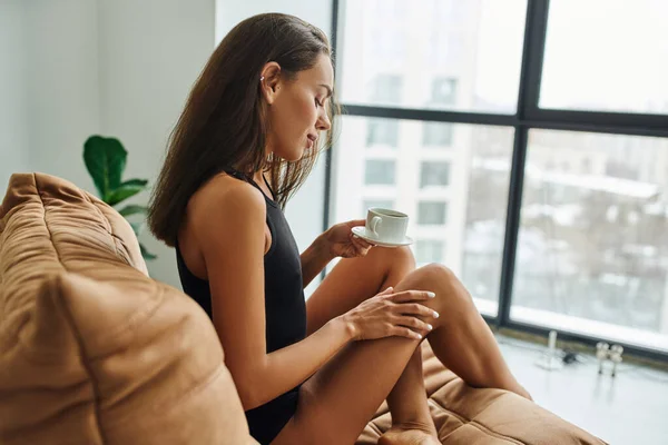 Seitenansicht, junge Frau mit brünetten Haaren, die eine Tasse Morgenkaffee in der Hand hält, sitzt auf einem Bohnensackstuhl — Stockfoto