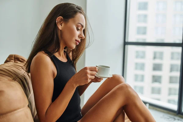 Brünette junge Frau mit langen Haaren, die eine Tasse Morgenkaffee in der Hand hält und auf einem Bohnensackstuhl sitzt — Stockfoto