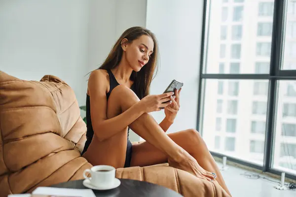 Hübsche Frau mit Smartphone und sitzt auf Bohnensackstuhl, Couchtisch mit verschwommener Tasse — Stockfoto