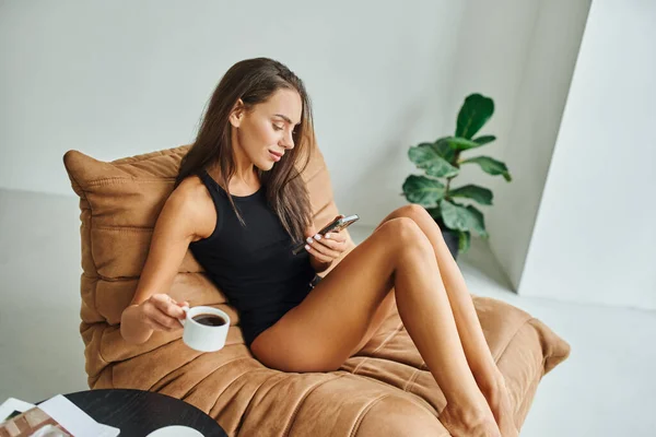 Mujer satisfecha usando el teléfono inteligente y sosteniendo la taza de café negro, sentado en la silla de la bolsa de frijol en casa - foto de stock