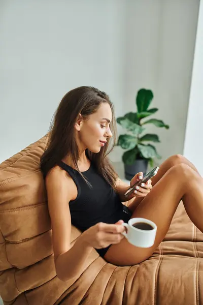 Femme heureuse en utilisant un smartphone et tenant une tasse de café noir, assis sur une chaise de sac de haricots à la maison — Photo de stock