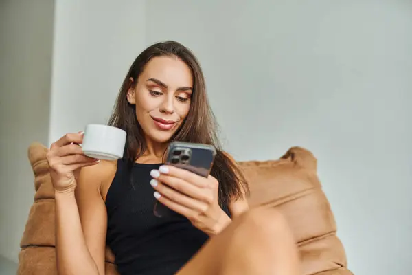 Mujer positiva usando teléfono inteligente y sosteniendo la taza de café, sentado en la silla de la bolsa de frijol en casa - foto de stock