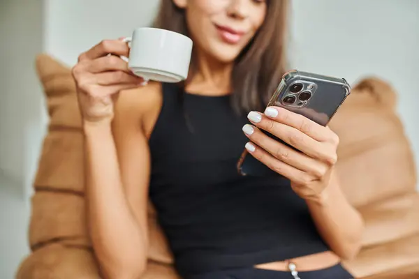 Обрізаний вид щасливої жінки за допомогою смартфона і тримає чашку кави вдома, бобовий мішок стілець — стокове фото