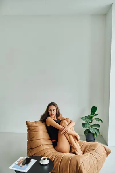 Attrayant pieds nus femme assise sur une chaise de sac de haricot près de la table basse avec tasse de café noir — Photo de stock