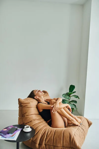 Attraente donna a piedi nudi poggiata sulla sedia sacchetto di fagioli vicino al tavolino con tazza di caffè nero — Foto stock