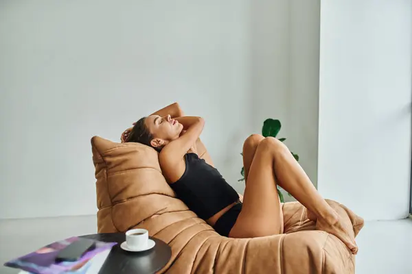 Belle femme pieds nus reposant sur une chaise de sac de haricot près de la table basse avec tasse de café noir — Photo de stock