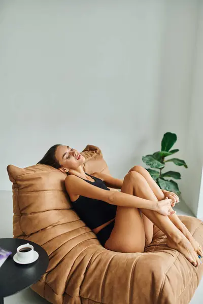 Hermosa mujer joven descansando en la silla de la bolsa de frijol cerca de la mesa de café con taza de café negro - foto de stock