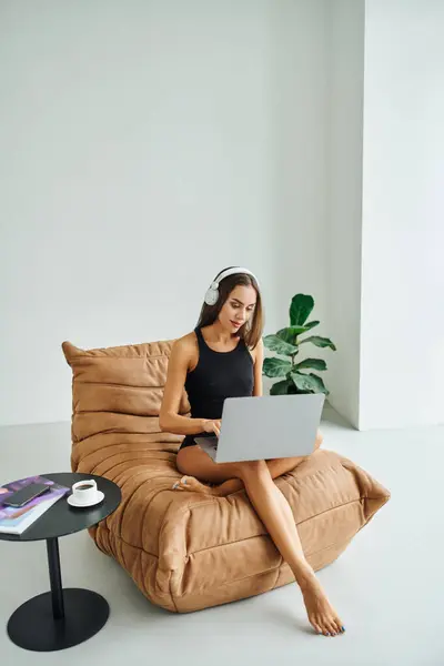 Bastante freelancer en auriculares inalámbricos utilizando el ordenador portátil y sentado en la silla de la bolsa de frijol, mujer joven - foto de stock