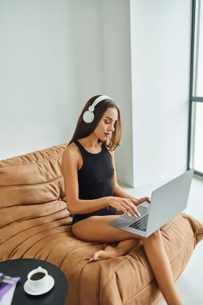 Freelancer descalço em fones de ouvido sem fio usando laptop e sentado em cadeira de saco de feijão, mulher bonita — Fotografia de Stock