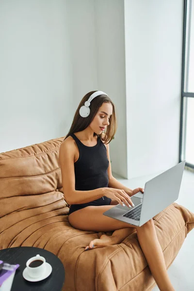 Freelance a piedi nudi in cuffie senza fili utilizzando il computer portatile e seduto sulla sedia sacchetto di fagioli, bella donna — Foto stock