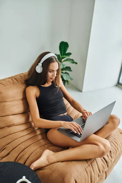 Bella freelance in cuffie wireless con computer portatile e seduto sulla sedia sacchetto di fagioli, giovane donna — Foto stock