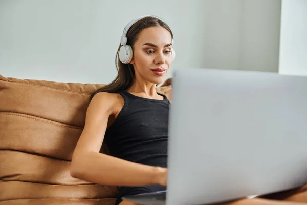 Mujer bonita en auriculares inalámbricos usando el ordenador portátil y sentado en la silla de la bolsa de frijol, trabajo remoto - foto de stock