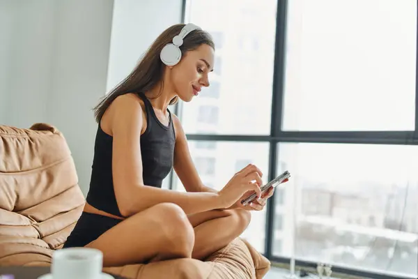 Pretty woman in wireless headphones using smartphone and sitting on bean bag chair, weekend vibes — Stock Photo