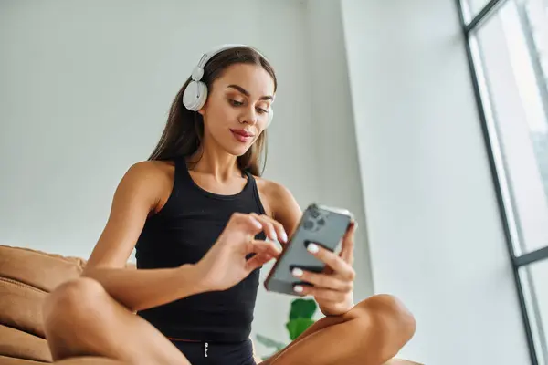 Mujer complacida en auriculares inalámbricos con teléfono inteligente y sentado en la silla de la bolsa de frijoles, vibraciones de fin de semana - foto de stock