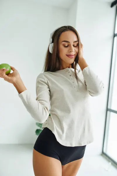 Femme joyeuse à manches longues et culotte écouter de la musique dans les écouteurs et tenant pomme — Photo de stock