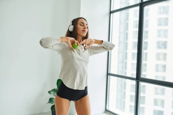 Emotional woman in long sleeve and panties listening music in headphones and holding apple — Stock Photo