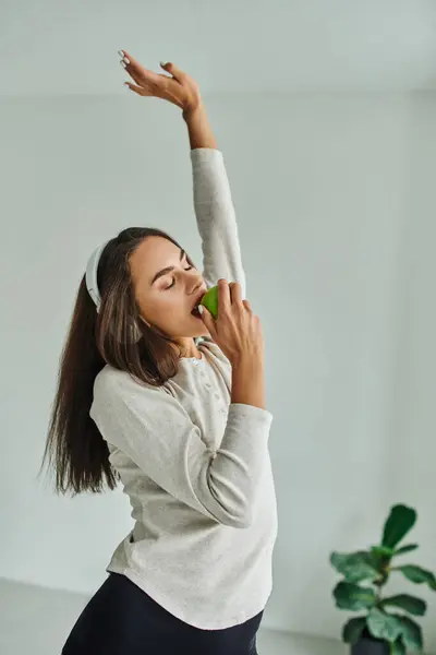 Brunette woman in long sleeve listening music in headphones and biting green apple, enjoying taste — Stock Photo