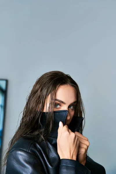 Femme brune aux cheveux mouillés et aux yeux bleus ajustant le col de manteau en cuir noir sur fond gris — Photo de stock