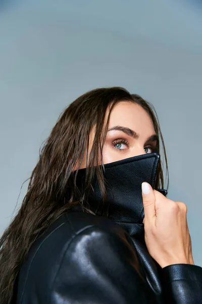 Beautiful woman with wet hair and blue eyes adjusting collar of black leather coat on grey backdrop — Stock Photo