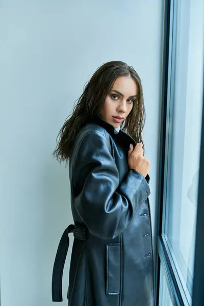 Pretty woman with brunette long hair posing in black leather coat near window at home, seductive — Stock Photo