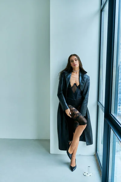 Donna appassionata con bruna capelli bagnati in posa in cappotto di pelle nera e biancheria intima di pizzo vicino alla finestra — Foto stock