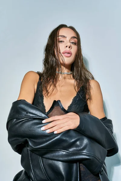 Donna sexy con capelli castani in posa in biancheria intima nera, corsetto di pizzo e cappotto di pelle su grigio — Foto stock