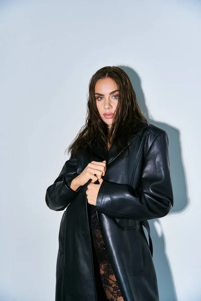 Сексуальная внешность, женщина с волосами брюнетки позирует в черном кружевном нижнем белье и кожаном пальто на сером заднем плане — стоковое фото