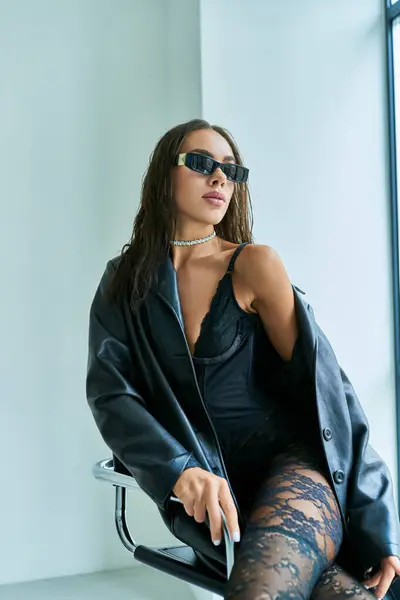Mulher sedutora em óculos de sol, roupa interior de renda preta e casaco de couro sentado na cadeira, fundo cinza — Fotografia de Stock