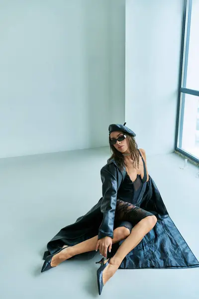 Ropa sexy, mujer seductora en boina, gafas de sol, ropa interior de encaje y abrigo de cuero sentado en el suelo - foto de stock