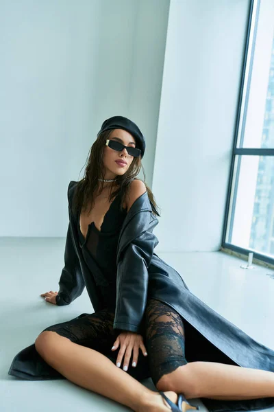 Sexy Outfit, schöne Frau mit Baskenmütze, Sonnenbrille, Spitzenunterwäsche und Ledermantel auf dem Boden sitzend — Stockfoto