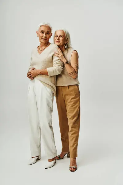 Elegantes mujeres mayores en elegante atuendo casual de pie en gris, el envejecimiento positivo de las amigas - foto de stock