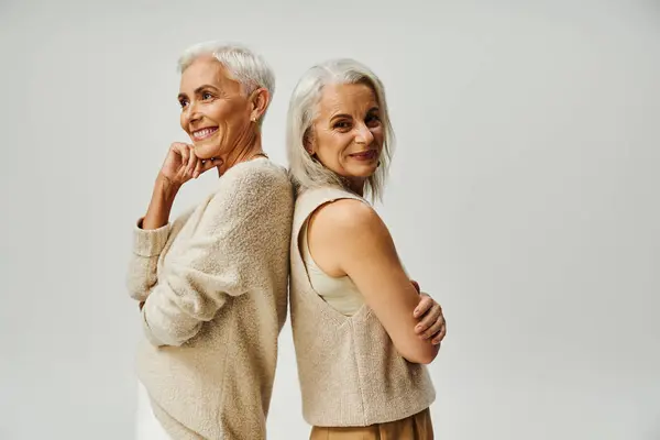 Stylische Seniorin lächelt in die Kamera und steht Rücken an Rücken mit einer glücklichen Freundin auf grau — Stockfoto