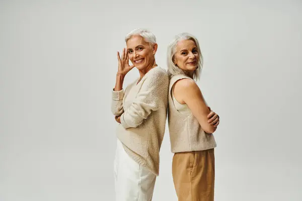 Envelhecimento graciosamente, mulheres seniores elegantes de pé de volta para trás e sorrindo para a câmera em cinza — Fotografia de Stock