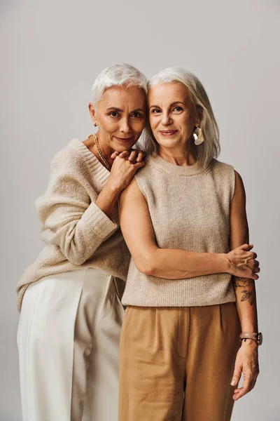 Mature woman leaning on shoulder of happy female friend looking at camera on grey, graceful aging — Stock Photo