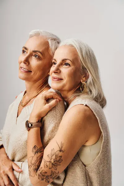 Dreamy and happy senior women in makeup and golden accessories looking away on grey, ageless beauty — Stock Photo