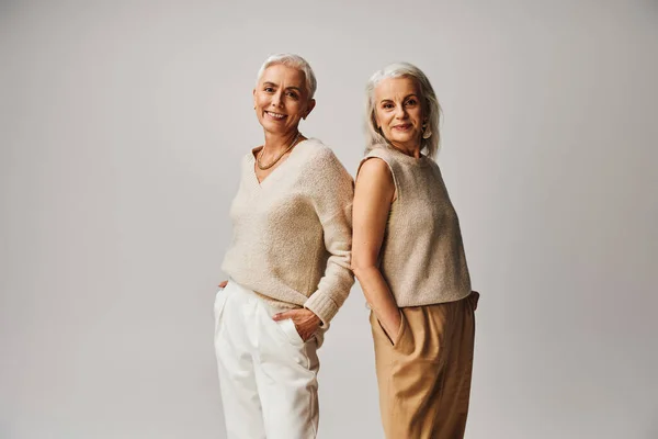 Lächelnde Seniorinnen in modischer Kleidung, Rücken an Rücken stehend, die Hände in den Taschen auf grau — Stockfoto
