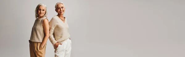 Счастливые старшие модели в модной одежде, позирующие спина к спине с руками в карманах на сером, баннер — стоковое фото