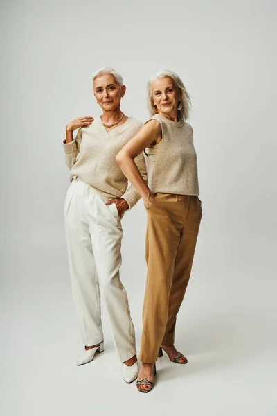 Longitud completa de elegantes fashionistas senior posando con las manos en bolsillos sobre fondo gris - foto de stock