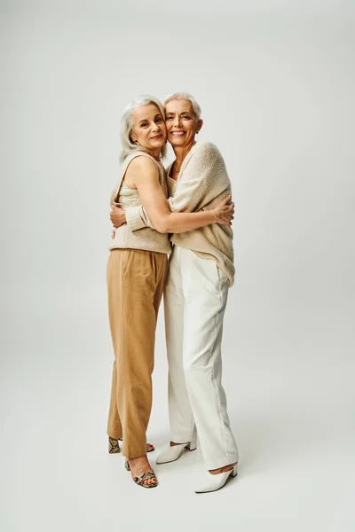 Full length of elegant and happy mature women in stylish pastel clothes hugging each other on grey — Stock Photo