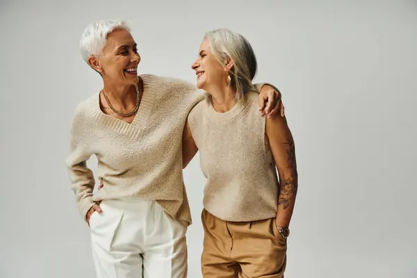 Fashionable senior female friends smiling at each other while posing with hands in pockets on grey — Stock Photo