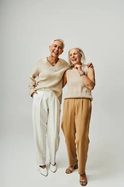 Modern and fashionable senior female friends embracing and laughing on grey background, full length — Stock Photo