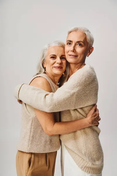 Alegres mujeres mayores en ropa pastel de moda abrazando en fondo gris, amistad de por vida - foto de stock