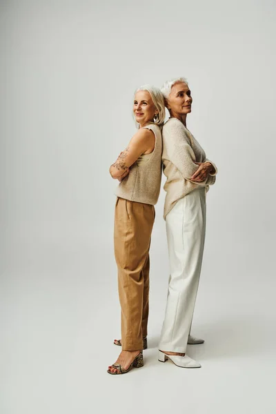Full length of senior fashionistas standing back to back with folded arms on grey, lifestyle friends — Stock Photo