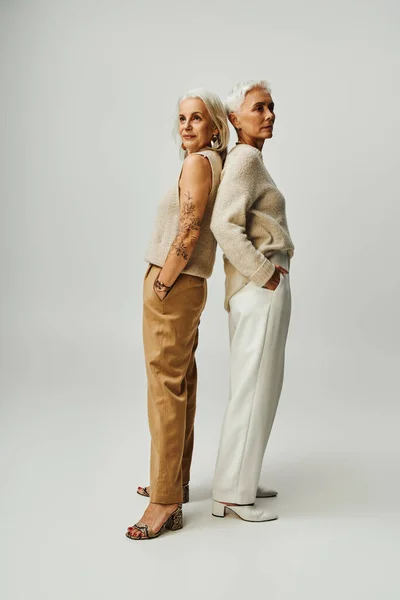 Volle Länge selbstbewusster Seniorinnen, die Rücken an Rücken mit den Händen in den Taschen vor grauem Hintergrund stehen — Stockfoto