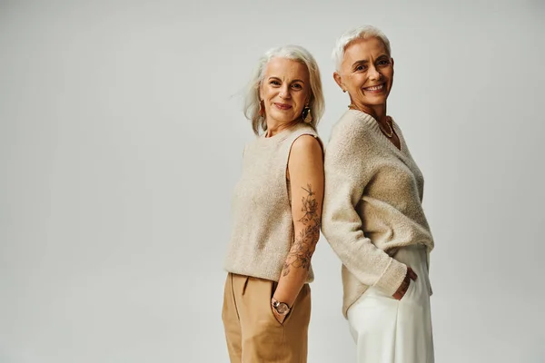 Happy mature ladies looking at camera while standing back to back on grey, fashionable seniors — Stock Photo