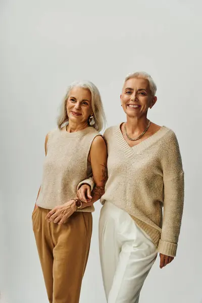 Pleased mature women in trendy casual clothes standing arm in arm on grey, fashion and friendship — Stock Photo