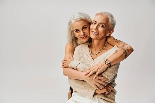 Mujer tatuada de moda abrazando madura encantada amiga en gris, estilo de vida positivo - foto de stock
