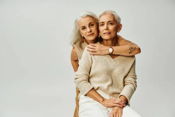 Гламурная татуированная женщина, обнимающая улыбающуюся и стильную подругу, сидящую на серых, модных пожилых людях — стоковое фото