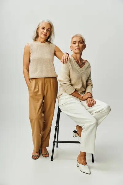 Eitelkeit fairen Stil, lächelnde Seniorin in der Nähe modische Freundin sitzt auf Stuhl auf grau — Stockfoto